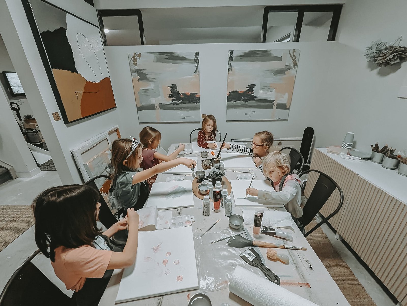 Wide view of a Painting Prodigies class at the studio table, two large paintings on the wall behind