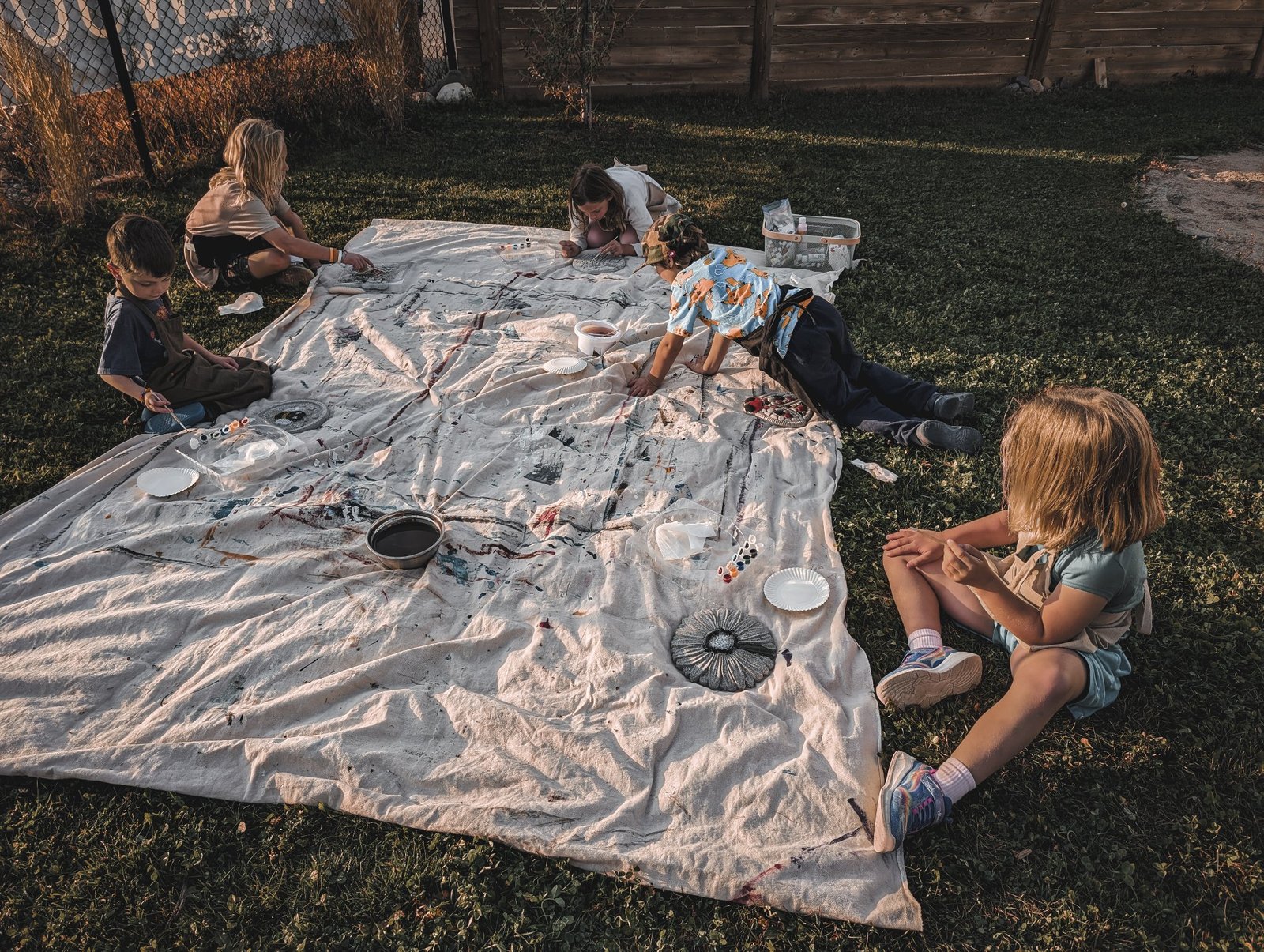 Children painting outdoors at golden hour on a paint-marked drop cloth on the grass, seen from above and behind