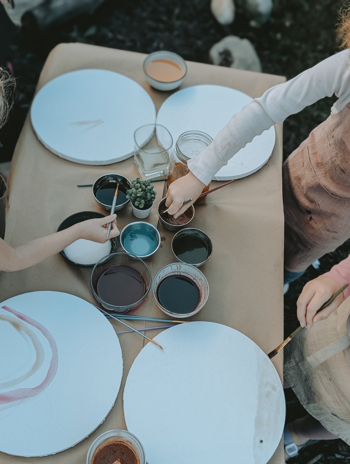 Overhead view of children's hands reaching for bowls of natural pigment beside round canvases at a Painting Prodigies class, no faces shown