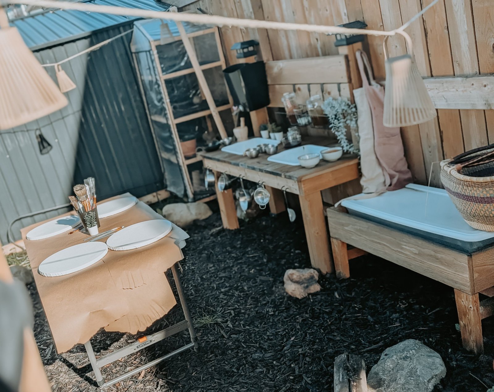 The outdoor studio: a wooden work station with round canvases on the table, a greenhouse and hanging lamps behind