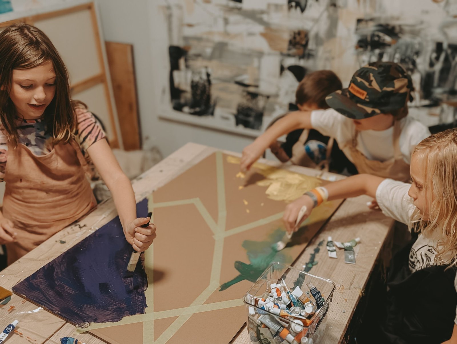 Four children painting a large taped geometric canvas together around the studio table at a Painting Prodigies class, faces turned to their work, with Leta Taylor's black, white, and gold artwork on the wall behind them